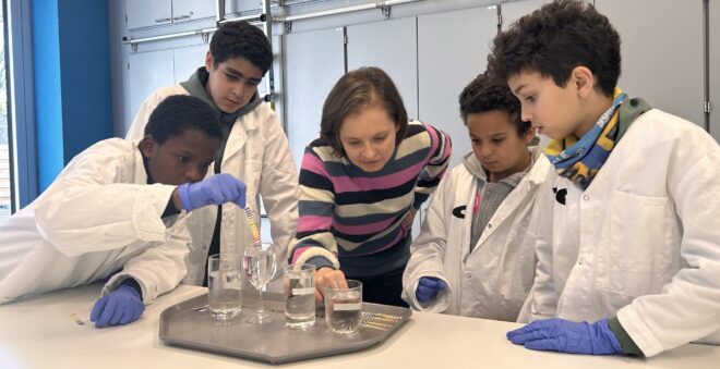 Galapagos STEM Ambassador Sara Musch is in the lab, explaining a lab test to a group of young children wearing lab coats and protective gloves